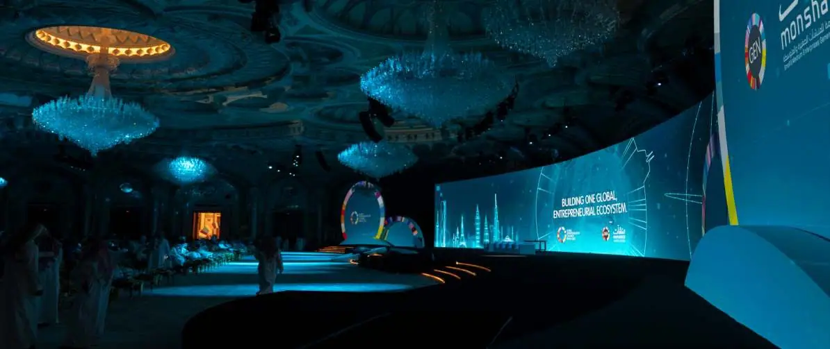 Wide view of the Global Entrepreneurship Congress main hall in Riyadh: chandeliers and a curved stage screen with GEN and Monsha'at branding, the audience far in the distance