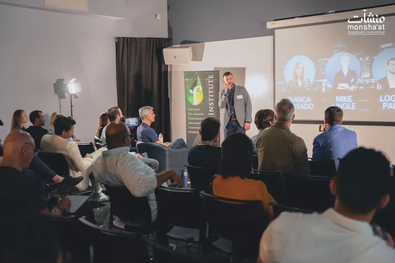 A speaker addresses an audience in a startup event space abroad, with speaker portraits projected on a screen